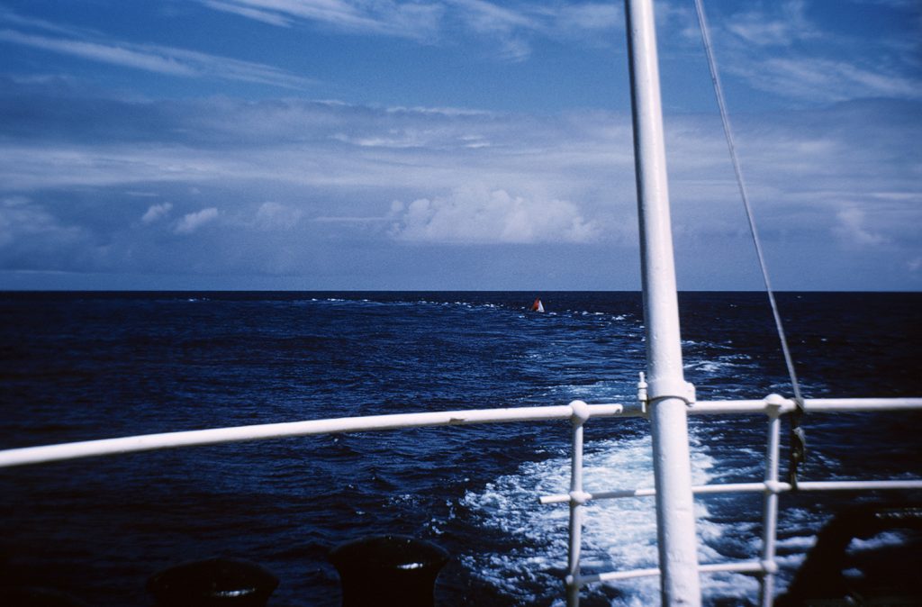 Looking over the stern of a ship out at sea, with a small red boat in it's wake